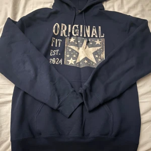 Mörkblå hoodie med stjärntryck - Säljer en mörkblå hoodie med tryck 'ORIGINAL FIT EST. 2024' och en stor stjärna omgiven av mindre stjärnor på bröstet. Tröjan har huva med dragsko och en stor magficka framtill. Perfekt för en avslappnad stil.