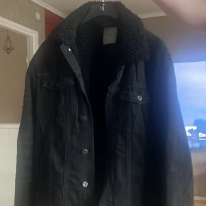 Svart jeansjacka med foder - Säljer en svart jeansjacka med mysigt teddyfoder i kragen. Klassisk design med knappar framtill och bröstfickor. Perfekt för kyligare dagar och passar till det mesta.