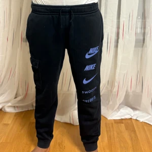 Svarta mjukisbyxor från Nike med tryck - Svarta mjukisbyxor från Nike med blå loggor och text på ena benet. Byxorna har resår i midjan med snörning, en cargoficka på sidan och muddar vid bensluten. Perfekta för en avslappnad stil. Knappt använd 