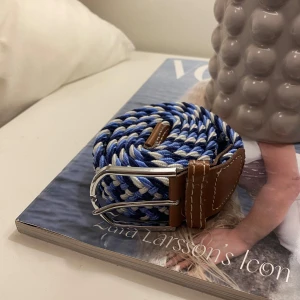 Blåvitt flätat bälte med bruna skinn-detaljer - Säljer ett snyggt flätat bälte i blått och vitt med bruna skinn-detaljer vid spännet. Bältet har ett silverfärgat metallspänne och passar perfekt till jeans eller chinos. Stilren och modern accessoar för att lyfta din outfit.