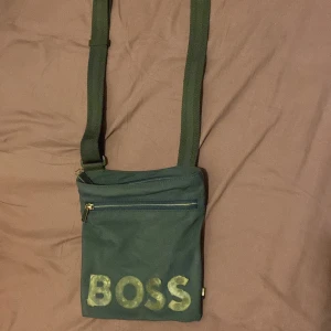 Mörkgul axelväska från BOSS - Säljer en mörkgult axelväska från BOSS med justerbar rem och dragkedja framtill. Väskan har tydlig BOSS-logga på framsidan och är perfekt för att bära med sig det viktigaste. Snygg och enkel design. Den är lite gammal men funkar säljer den för bra pris