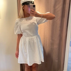 Vit kort klänning med puffärmar och broderade blommor - Superfin vit klänning med puffärmar och broderade blommor. Klänningen har öppen rygg med knytband och en söt, luftig känsla. Perfekt till student eller sommarens festligheter. Kort modell med rund halsringning och markerad midja.🎓🌸💗 helt ny med prislapp kvar