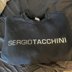 Svart sweatshirt från Sergio Tacchini - Svart sweatshirt från Sergio Tacchini med stor vit logga framtill och tryckt emblem på ärmen. Klassisk rund halsringning och normal passform. Perfekt för en sportig och avslappnad stil.