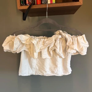 Offshoulder volangtopp från Gina Tricot - Säljer en ljusbeige offshoulder topp från Gina Tricot med volangdetaljer runt hela överdelen. Toppen är croppad och har dragkedja i sidan för enkel påklädning. Storlek 38 men passar xs/s/m