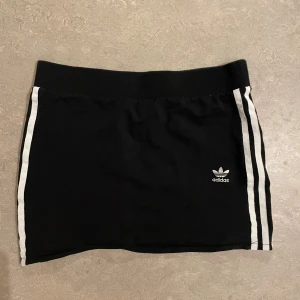 Svart kjol med vita ränder från Adidas - Svart kortkjol från Adidas med klassiska vita ränder längs sidorna och Adidas-logga framtill. Bred resår i midjan för en bekväm passform. Sportig och stilren design som passar perfekt till vardags eller träning.