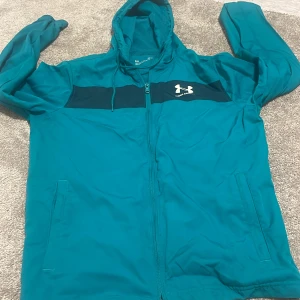 Turkos vindjacka från Under Armour - Säljer en turkos vindjacka från Under Armour med svart panel över bröstet och vit logga. Jackan har huva, dragkedja framtill och två sidofickor. Perfekt för träning eller utomhusaktiviteter under vår och höst.