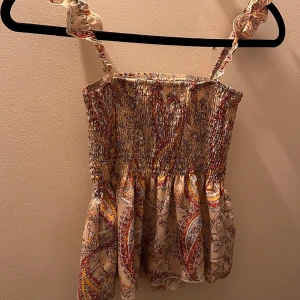 Sommar topp - Säljer en beige linnetopp med färgglatt paisleymönster i rött, gult och svart. Toppen har smock över bysten och volangiga axelband. Perfekt för varma dagar och ger en bohemisk känsla.💞