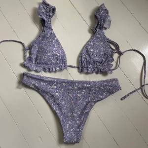 Ljuslila blommig bikini med volangdetaljer - Superfin ljuslila bikini med små vita blommor och volangdetaljer på både överdel och underdel. Bikinin har knytband och en feminin, söt design som passar perfekt till sommaren. Aldrig använd men alla lappar är borta. Skicka för fler bilder💞
