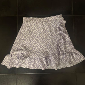 Ljuslila blommig kjol med volang - Supersöt ljuslila kjol med små vita blommor och volangdetalj längs nederkanten. Kjolen har en lätt är perfekt för en somrig dag. Bekväm passform och luftig känsla.