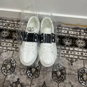 Valentino open  - Säljer ett par vita sneakers från Valentino Garavani med ett svart band och vita nitar över vristen. Skorna har rund tå, platt sula och snörning. Märkeslogga i guld på plösen. Perfekta för dig som gillar stilrena och exklusiva sneakers.Helt oanvända med ingen box tyvärr