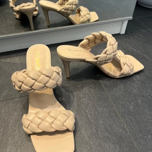 Beige flätade klack-sandaler - Säljer ett par stilrena beige sandaler med flätade remmar och hög klack. Skorna har fyrkantig tå och två breda, vadderade band över foten. Perfekta för dig som vill ha en trendig och elegant look.