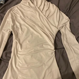 Beige omlottopp med lång ärm - Snygg beige omlottopp med lång ärm och draperad detalj vid axeln. Toppen har en elegant omlott-skärning och är tillverkad i ett mjukt, stretchigt material. Perfekt för dig som gillar stilrena och trendiga plagg.