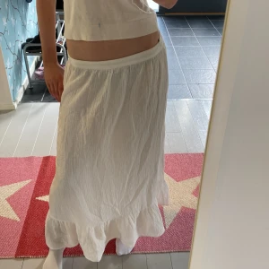 Vit långkjol med volang - Säljer en vit långkjol med bred resår i midjan och volang längst ner. Kjolen har en luftig och flowy känsla, perfekt för varma dagar. Enkel att matcha med olika toppar för en avslappnad stil. Storlek M men passar även S