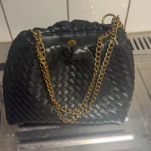 Svart flätad axelväska med guldkedja - Säljer en svart axelväska med flätad design och snygg guldfärgad kedja som axelrem. Väskan har ett klassiskt och stilrent utseende med tryckknappsstängning och innerfack. Perfekt för dig som vill ha en trendig accessoar. Helt nytt skick 