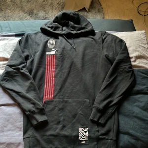 Svart hoodie och byxa från Puma x AC Milan - Svart hoodie och byxa från Puma i samarbete med AC Milan. Tröjan har huva med dragsko, magficka och röda vertikala ränder på framsidan samt AC Milan-logga och Puma-logga på bröstet. Ribbstickade muddar vid ärmslut och nederkant.