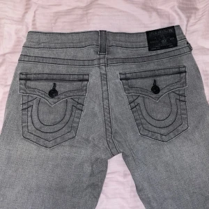 True religion - Säljer ett par grå lågmidjade jeans från True Religion med klassiska bakfickor och svarta knappar. Byxorna har normal passform och är ”straight”. Snygga detaljer med kontrastsömmar och logga på bakfickan. Helt oanvända. 