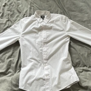 Vit skjorta från H&M - Klassisk vit skjorta från H&M med lång ärm och knappar hela vägen framtill. Skjortan har en slim fit och är tillverkad i bomull, perfekt för en clean och stilren look.