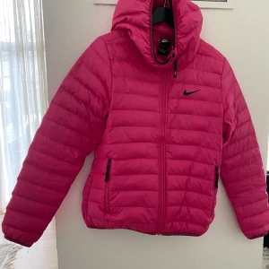 Rosa pufferjacka från Nike - Säljer en snygg rosa pufferjacka från Nike med huva och svarta dragkedjor. Jackan har quiltad design och Nike-logga på bröstet. Perfekt för kyliga dagar när du vill sticka ut lite extra.