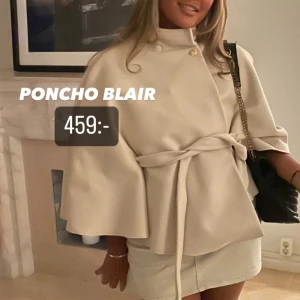 Beige poncho från missäy - Poncho i beige med hög krage, knappar vid axeln och knytskärp i midjan. Modellen har vida ärmar och en elegant, lös passform. Perfekt för lager-på-lager och stilrena outfits.