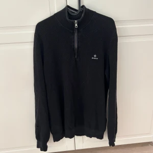 Quarter Zip från Gant - Litet hål som går att sy på armen. Svart stickad tröja från Gant med halv dragkedja och liten logotyp på bröstet. Tröjan har lång ärm och ribbade muddar vid ärmslut och nederkant. Perfekt för lager-på-lager under kyliga dagar.