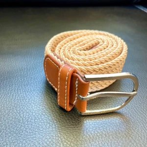 Beige flätat bälte med bruna skinn-detaljer - Säljer ett snyggt beige flätat bälte med bruna skinn-detaljer och silverspänne. Bältet har en stilren design och passar perfekt till jeans eller chinos. Flätningen gör det enkelt att justera längden efter behov.