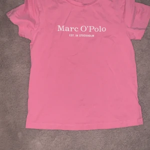 Rosa t-shirt från Marc O'Polo - Aldrig använt, säljer en rosa t-shirt från Marc O'Polo med vit logga och texten 'EST. IN STOCKHOLM' framtill. Klassisk passform med rund hals och korta ärmar. Perfekt för en enkel och stilren look.
