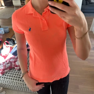 Orange pikétröja från Ralph Lauren - Säljer en orange pikétröja från Ralph Lauren med klassisk krage och korta ärmar. Tröjan har blå broderad logga på bröstet och är i ett mjukt material