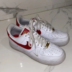 Nike Air Force 1 med röd swoosh och gulddetaljer - Säljer ett par klassiska Nike Air Force 1 i vitt med röd swoosh och små gulddetaljer. Skorna har snörning och är i syntetmaterial med rund tå och platt sula. Perfekta för dig som gillar en sportig och stilren look.