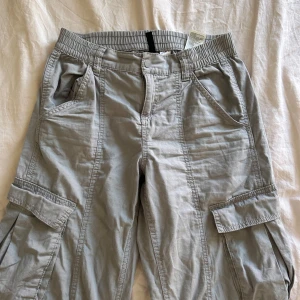 Ljusgrå cargopants med fickor - Säljer ett par ljusgrå cargopants med flera stora fickor på sidorna och resår i midjan. Byxorna har raka ben och klassisk cargostil, perfekta för en avslappnad look.