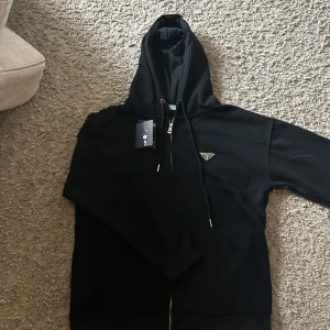 Prada zip hoodie - Säljer en svart hoodie från Prada med dragkedja och huva. Tröjan har en klassisk passform och är dekorerad med Pradas ikoniska metallogo på bröstet. Perfekt för dig som vill ha en stilren och lyxig look.Storlek S men större i storleken