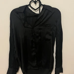 Svart satinblus från Gina Tricot - Säljer en svart, glansig blus från Gina Tricot i satinmaterial. Blusen har lång ärm, klassisk krage och knäppning framtill. Perfekt för dig som gillar en stilren och elegant look.