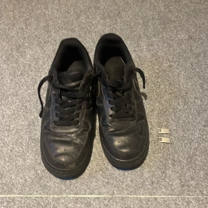 Svarta Nike Air Force 1 sneakers - Klassiska helsvarta Nike Air Force 1 sneakers med svarta skosnören och rund tå. Skorna har en platt sula och medföljer två metalltaggar med 'AF1'-text. Perfekta för en stilren och enkel look.pris kan diskuteras 