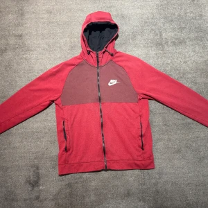 Röd hoodiejacka från Nike med dragkedja - Röd hoodiejacka från Nike med svart innerfoder i huvan och vit logga på bröstet. Jackan har hel dragkedja framtill, två fickor med dragkedja och lite sliten logga vid ärmslutet. Perfekt för en sportig stil.