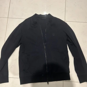 Svart zip-jacka från J.Lindeberg - Säljer en svart zip-jacka från J.Lindeberg med diskret logga på bröstet. Jackan har lång ärm, två fickor framtill och ribbade muddar. Enkel och stilren design som passar till många tillfällen.