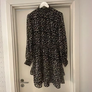 Svart blommig kortklänning med volang - Kortklänning med svart bas och vitt blommönster. Hög hals, långa ärmar med resår vid handlederna och volangdetaljer i kjolen. Perfekt för dig som gillar en feminin och trendig stil.