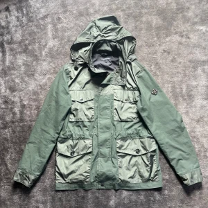 Grön vindjacka från J.Lindeberg - Snygg grön vindjacka från J.Lindeberg med huva i nacken plus flera praktiska fickor och en patch på ena ärmen. Perfekt för blåsiga dagar. Storlek M 