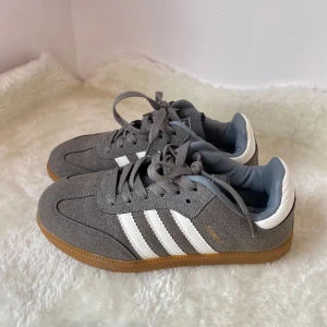 Grå Adidas Samba sneakers - Säljer ett par grå Adidas Samba sneakers med vita ränder och detaljer samt gummisula. Skorna har klassisk design med snörning och är tillverkade i mocka och syntet. Perfekta för dig som gillar stilrena och sportiga skor. Aldrig använda!