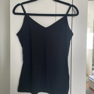 Svart ribbad linnetopp - Säljer en svart ribbad linnetopp med smala axelband och v-ringning. Toppen har en enkel och stilren design som passar perfekt till sommaren. Materialet är mjukt och följsamt.