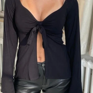 Svart långärmad topp med knyt från DesignSi - Säljer en svart långärmad topp från DesignSi med djup v-ringning och knyt framtill. Toppen har en figurnära passform och är tillverkad i ett mjukt, stretchigt material. Perfekt att styla med jeans eller kjol. Använd några gånger 