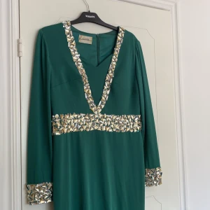 Grön långklänning med paljett-detaljer - Säljer en elegant grön långklänning med djup v-ringning och glittriga paljett-detaljer längs halsringning, midja och ärmslut. Klänningen har lång ärm och en slits framtill för extra stilpoäng. Perfekt för dig som vill sticka ut! Använd en gång!