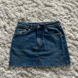 Jeanskjol från Zara - Säljer en klassisk blå jeanskjol från Zara med rå kant nertill och femficksmodell. Kjolen har normal passform och är i kort modell, perfekt till sommaren. Stängs med knapp och dragkedja framtill.