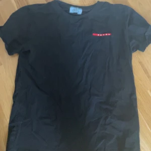 Svart t-shirt från Prada - Svart t-shirt från Prada med liten röd logotyp på bröstet. Klassisk passform och rund halsringning. Perfekt för en stilren och enkel look.