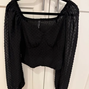 Svart blus med prickig struktur från new yorker - Säljer en svart blus från new yorker med långärmade transparenta ärmar och prickig struktur. Blusen har croppad passform, knappar framtill och smockad rygg för extra komfort. Perfekt för dig som gillar trendiga detaljer!