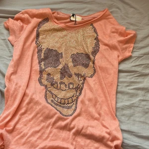 Persikofärgad t-shirt med döskalle från Chica London - Säljer en supercool persikofärgad/rosa  t-shirt från Chica London med ett stort döskallemotiv i glittriga nitar på framsidan. T-shirten har korta ärmar och en avslappnad passform. Den är tyvärr lite sliten längst ner i sömmen som man ser på sista bilden men den är köpt från sellpy och aldrig använd av mig💓 
