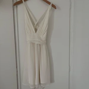 Vit omlottklänning med v-ringning - Säljer en vit kortklänning med djup v-ringning och omlottdetalj i midjan. Klänningen har smala axelband som korsas i ryggen och är ärmlös. Tyget är lätt och faller fint, perfekt för sommarens tillfällen.