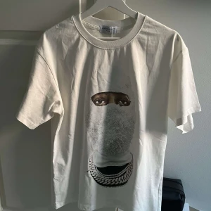 Vit t-shirt med tryck från Ih Nom Uh Nit - Säljer en vit t-shirt från Ih Nom Uh Nit Paris med ett stort tryck av ett ansikte med mask och kedja på framsidan. T-shirten har rund hals och normal passform. Perfekt för dig som gillar streetwear och unika prints. Passar även M. Den är stor i storlek 