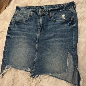 Jeanskjol med slits och fransad kant - Säljer en blå jeanskjol med rå fransad nederkant och slitningar fram. Kjolen har klassiska fickor både fram och bak samt en tydlig slits framtill. Perfekt för dig som gillar en lite tuffare stil.