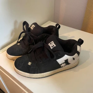 Svarta DC sneakers med vita detaljer - Säljer ett par klassiska svarta DC sneakers med vita detaljer och logga på sidan. Skorna har snörning, platt sula och rund tå. Perfekta för skate eller vardagsstil. Märket är tydligt på både plös och sula.