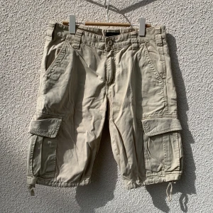 Beiga cargoshorts från Gant - Säljer ett par beiga cargoshorts i storlek 32 från Gant med flera fickor och snörning vid benslut. Shortsen har normal passform och är tillverkade i 100% bomull.