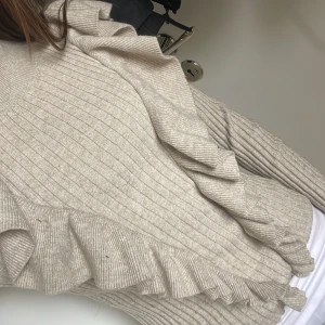 Beige ribbad tröja med volang - Säljer en beige ribbad tröja med volangdetaljer längs framsidan och ärmarna. Tröjan har rund halsringning och långa ärmar. Perfekt för dig som gillar en trendig och feminin stil.